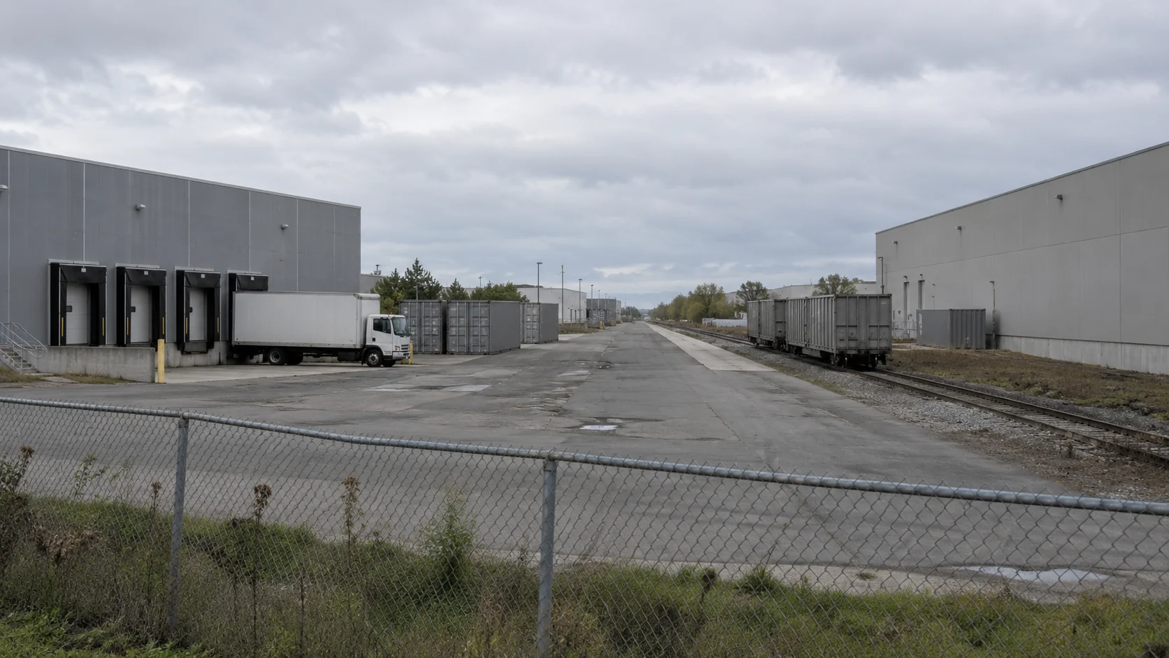 Warehouses beside a freight access road, with a plain white truck and grey rail wagons behind a wire fence